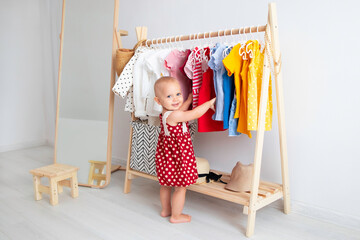 baby girl stands near a wardrobe and chooses a dress and smiles. dressing closet with clothes arrang