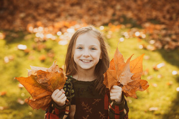 Funny kid outdoors in autumn park. Adorable happy girl playing with fallen leaves in autumn park. Beautiful happy girl having fun in autumn park among fallen leaves.