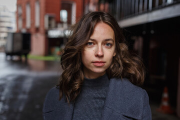 young thoughtful pretty girl in gray coat and turtleneck poses in front of red building. Street style portrait photo session of attractive female, urban photoshoot of beautiful elegant model