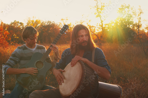 Fotografía Two hippie men playing djembe drum and acoustic guitar in the autumn in the forest at sunset