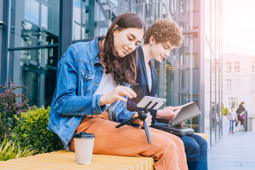 Two different gender male and female students friends sitting on the bench outdoor, using smart phone, working together, discuss joint project, relaxing during the break, drinking coffee, reading book