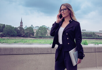 Young career woman, talks on the phone and holds documents and her laptop. In the background, the river is a city.