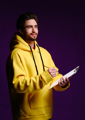 Wall Mural - A young man of 25-30 years in glasses and a yellow sweatshirt holding a clipboard with clip in one hand and a pencil in the other