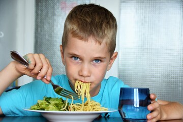 boy with blue eyes in a blue T-shirt eats spaghetti and holds a glass of clean water
