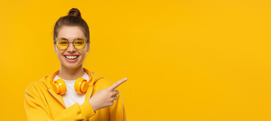 banner of young laughing girl wearing headphones around neck, pointing right, isolated on yellow bac