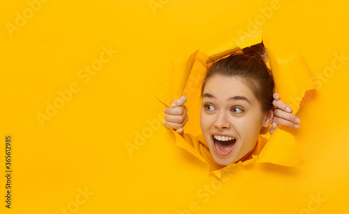 Fotomural Excited girl looking through hole in paper at left with eyes round with surprise