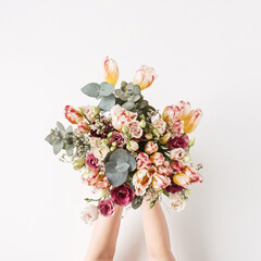 Wall Mural - Female hand holding tulip, eucalyptus flowers bouquet against white wall. Holiday celebration festive floral concept