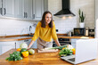 © Vadim Pastuh - A young woman in yellow shirt cooks in the kitchen and looks with interest at the laptop screen. Woman prepare veggie dish, vegetables on the table