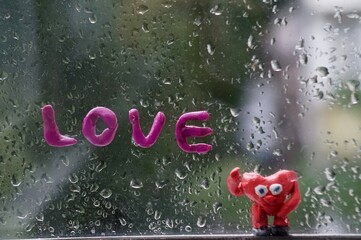 Sticker - A red heart made of plasticine against a wet window. Next to it is the inscription love.