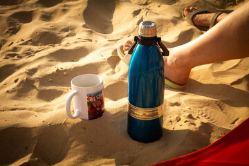 Thermos and a mug are standing on the sea sand in the sunset. A girl sits on a beach chair in shales.