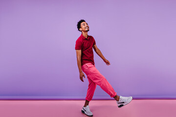 Wall Mural - Handsome african guy in trendy pants looking up with smile. Indoor photo of interested black male model dancing on purple background.