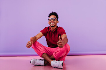 Wall Mural - Blithesome african man in sport shoes sitting on purple background. Chilling short-haired boy posing with thumb up.