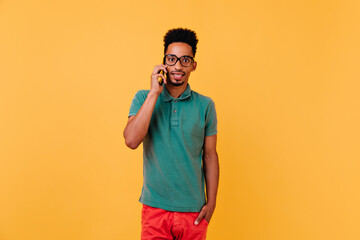 Wall Mural - Surprised black male model talking on phone. Studio shot of stylish african guy in glasses posing on yellow background with smartphone.