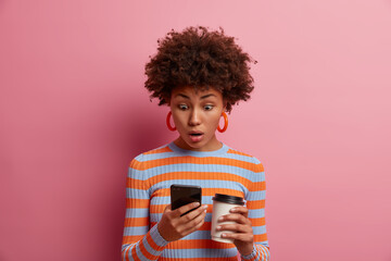 Wall Mural - Astonished surprised curly young woman stares at smart phone display, sees something amazing online, reads disturbing insulting message, drinks takeout coffee, poses against pink background.