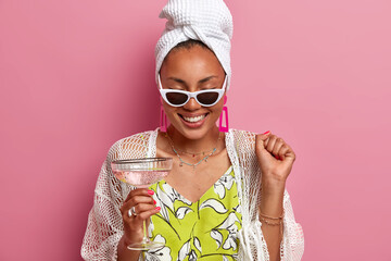 Wall Mural - Cheerful housewife has fun on pajama party, raises clenched fist, celebrates special event, drinks cocktail, wears bath towel on head, sunglasses, giggles positively, isolated over pink background