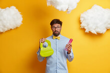 Stunned Bearded Father Busy Caring About Newborn, Going To Feed Baby, Holds Rubber Bib And Bottle With Nipple, Shocked As Wife Left Him With Child Alone, Poses Over Yellow Wall. Fatherhood Concept