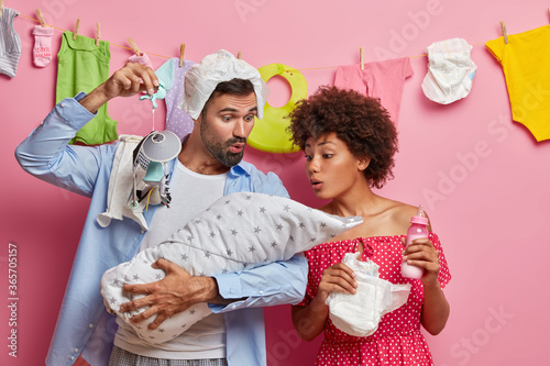 Caring young father and mother play and feed their little infant child, pose with newborn baby wrapped in blanket, show mobile and try to smooth, busy with parent chores. Happy family concept