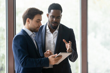 two multicultural male colleagues businessmen standing in modern office hall using device holding ta