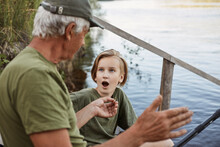 Grandfather And Grandson Fishing On River Berth, Senior Man Showing Size Of Fish He Caught Last Time, Male Child Posing With Opened Mouth, Being Shocked.