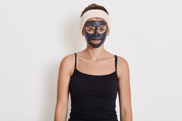 Beautiful girl with clay mask on her face standing with upset facial expression, looking at camera with sadness, wearing black t shirt and hairband.