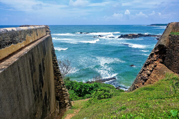 Ancient Fort Galle, Sri Lanka. Contrast between stone dilapidated walls and turquoise ocean. Summer sunny day, blue sky, foam on the waves. There are small rocky islands in the water. 