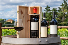 Three Bottles Including A Magnum In The Wooden Box And A Glass Of Red Wine On A Wooden Barrel With The Tuscan Countryside In The Background, Italy, On A Sunny Day
