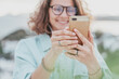 © olezzo - Beautiful happy curly young girl in glasses joyfully looks at the smartphone screen with a view of the city. Modern technologies and urban lifestyle