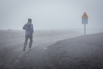 A man is running along the foggy road with blue speed limit and yellow windy road signs. Man is running to his 30s. Concept.