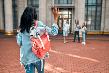 Wall Mural - A cute african american girl student with a pink backpack waving to a group of students near the campus.