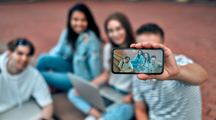 Wall Mural - A group of students are sitting on the steps near the campus with laptops, relaxing, chatting and taking selfies on a smartphone.