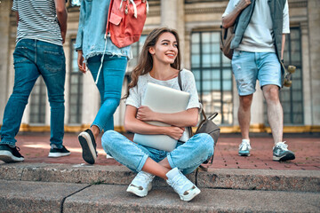 A charming girl student with a backpack and a laptop sits on the steps near the campus against the background of students passing by.