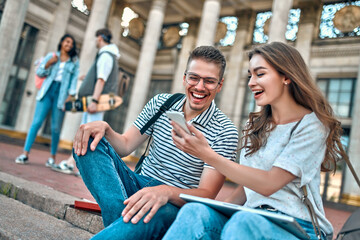 Wall Mural - A couple of students with backpacks and a laptop sit on the steps near the campus. The girl shows the guy something on her smartphone.