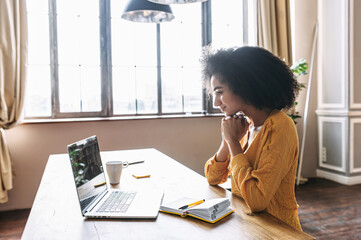 Distance communication with friends via video call. A young attractive mixed-race woman talking via video with her male friend, she looks at laptop screen with a smile. Side view