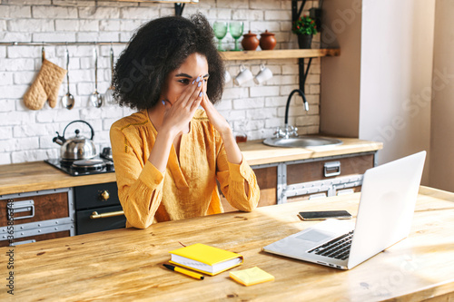 Unhappy mixed-race woman freelancer has a deadline with a remote work. Tired woman does not have time to finish on time, she sits in home office and covers eyes with hands