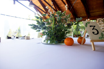 Composition of field herbs and cereals on the table in the restaurant in a transparent vase. White tablecloth and two tangerines. Stillife.