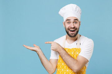 Excited young bearded male chef or cook baker man in apron white t-shirt toque chefs hat isolated on blue background studio. Cooking food concept. Mock up copy space. Pointing index finger hand aside.