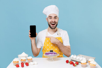 Wall Mural - Excited male chef or cook baker man in apron white t-shirt toque chefs hat cooking at table isolated on blue background. Cooking food concept. Pointing finger on mobile phone with blank empty screen.