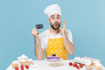 Wall Mural - Shocked young bearded male chef or cook baker man in apron white t-shirt toque chefs hat cooking at table isolated on blue background. Cooking food concept. Hold credit bank card, put hand on cheek.