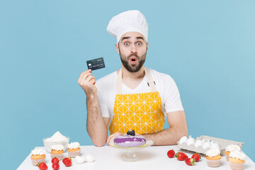 Wall Mural - Shocked young bearded male chef or cook baker man in apron white t-shirt toque chefs hat cooking at table isolated on blue background. Cooking food concept. Mock up copy space. Hold credit bank card.