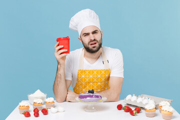 Wall Mural - Tired young bearded male chef or cook baker man in apron white t-shirt toque chefs hat cooking at table isolated on blue background in studio. Cooking food concept. Hold paper cup of coffee or tea.