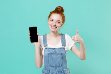 Smiling young readhead girl in casual denim clothes isolated on blue turquoise background . People lifestyle concept. Mock up copy space. Hold mobile phone with blank empty screen, showing thumb up.