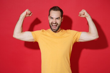 Strong funny young bearded man guy in casual yellow t-shirt posing isolated on red background studio portrait. People sincere emotions lifestyle concept. Mock up copy space. Showing biceps, muscles.