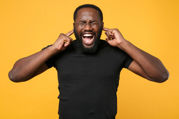 Wall Mural - Crazy african american man football fan in casual black t-shirt isolated on yellow background. People lifestyle concept. Mock up copy space. Covering ears with fingers, keeping eyes closed, screaming.