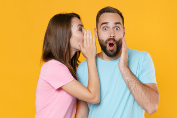 Shocked young couple friends guy girl in blue pink t-shirts isolated on yellow background. People lifestyle concept. Mock up copy space. Whispering secret behind hand, sharing news, put hand on cheek.