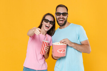 Wall Mural - Laughing couple two friends guy girl in t-shirts 3d glasses isolated on yellow background. People in cinema, lifestyle concept. Watching movie film hold bucket popcorn cup soda point finger on camera.