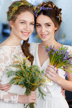 Vertical Portrait Of Two Brides In White Wedding Dresses With Flowers In Their Hair, With A Bouquet Of Wild Flowers On A Studio.