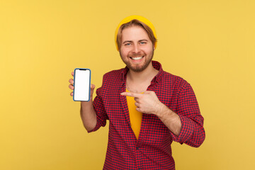 Wall Mural - happy trendy hipster guy in beanie hat and checkered shirt pointing mobile phone empty display, look