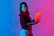 © khosrork - Neon light portrait of successful joyful office worker girl looking away with happy pensive face, smiling holding laptop, typing on keyboard, doing freelance job. colorful neon light, studio shot