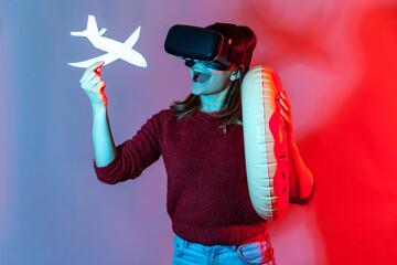 Joyful tourist girl in VR glasses holding rubber ring and airplane mock up, watching virtual travel tours, vacation in tropical country, having journey in augmented reality. neon light studio shot