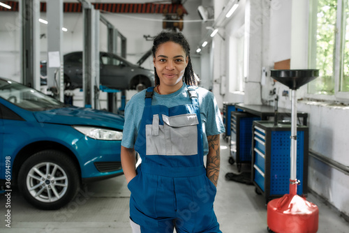 Come for help. Portrait of young african american woman, professional female mechanic in uniform smiling at camera, standing in auto repair shop. Car service, repair, maintenance and people concept